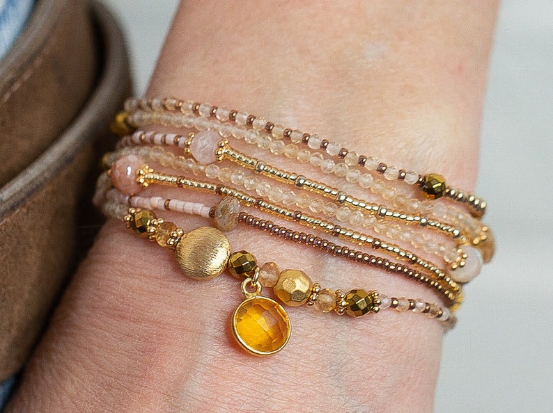 May include: A stack of delicate bracelets featuring a mix of gold, brown, and pink beads, with a gold charm and a yellow gemstone pendant.