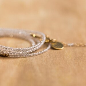 K&ouml;nnte beinhalten: Ein zartes, mehrstr&auml;ngiges Armband mit klaren, facettierten Perlen. Das Armband hat goldfarbene Akzente, darunter ein kleiner Anh&auml;nger und ein Verschluss. Der Schmuck wird auf einer hellbraunen Holzoberfl&auml;che pr&auml;sentiert.