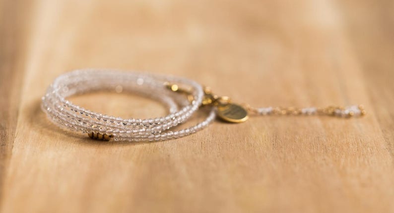 K&ouml;nnte beinhalten: Ein zartes, mehrstr&auml;ngiges Armband mit klaren, facettierten Perlen. Das Armband hat goldfarbene Akzente, darunter ein kleiner Anh&auml;nger und ein Verschluss. Der Schmuck wird auf einer hellbraunen Holzoberfl&auml;che pr&auml;sentiert.