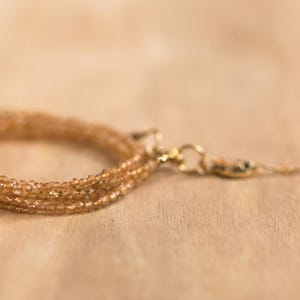 Op de afbeelding: Een delicate, meerstrengs armband in amberkleuren, met kleine, gefacetteerde kralen. De armband heeft een goudkleurige sluiting en een bungelende streng met een kleine kraal aan het uiteinde. De sieraden worden getoond op een lichtbruin houten oppervlak. Lengte armband: ca. 18 cm.
