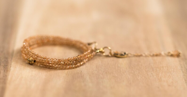 Op de afbeelding: Een delicate, meerstrengs armband in amberkleuren, met kleine, gefacetteerde kralen. De armband heeft een goudkleurige sluiting en een bungelende streng met een kleine kraal aan het uiteinde. De sieraden worden getoond op een lichtbruin houten oppervlak. Lengte armband: ca. 18 cm.