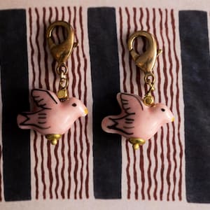 Puede incluir: Dos abalorios de cerámica rosa en forma de pájaro con detalles dorados y un cierre de langosta dorado. Los abalorios cuelgan de un fondo a rayas.