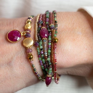 Puede incluir: Un conjunto de pulseras con cuentas rosas, verdes y doradas. Las pulseras presentan una variedad de cuentas de piedras preciosas, incluyendo rub&iacute; y esmeralda. Un dije dorado con una piedra de rub&iacute; cuelga de la parte superior de la pila.