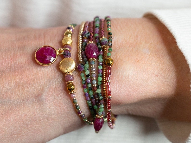 Puede incluir: Un conjunto de pulseras con cuentas rosas, verdes y doradas. Las pulseras presentan una variedad de cuentas de piedras preciosas, incluyendo rub&iacute; y esmeralda. Un dije dorado con una piedra de rub&iacute; cuelga de la parte superior de la pila.