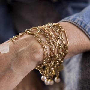 May include: A stack of gold chain bracelets with a white stone charm and pearl accents.