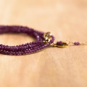 May include: A multi-strand bracelet with small, faceted, deep purple beads. The bracelet has a gold-coloured clasp and a delicate chain with matching beads extending from the clasp. The jewellery is displayed on a light brown wooden surface.