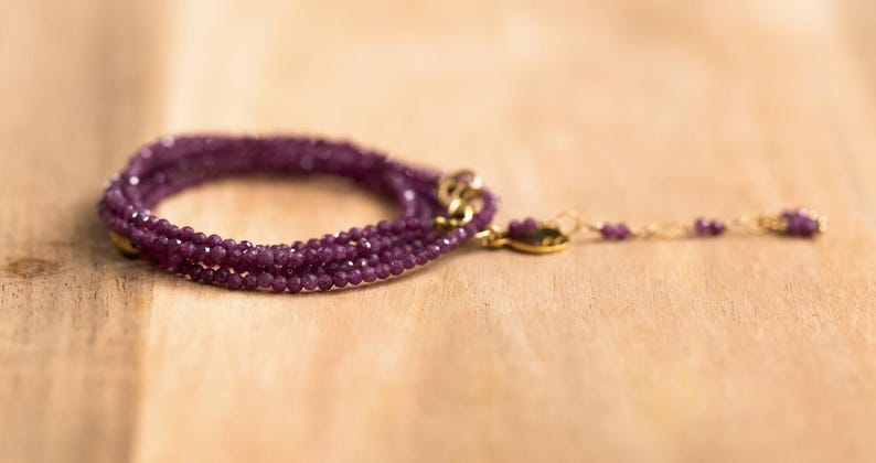 May include: A multi-strand bracelet with small, faceted, deep purple beads. The bracelet has a gold-coloured clasp and a delicate chain with matching beads extending from the clasp. The jewellery is displayed on a light brown wooden surface.