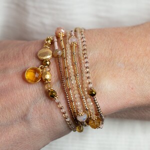 May include: A set of seven beaded bracelets in shades of beige, gold, and pink. The bracelets are stacked on a person's wrist and feature small gold and amber beads.