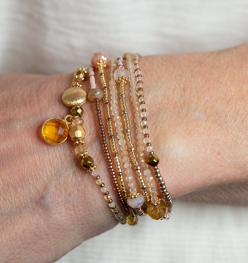 May include: A set of seven beaded bracelets in shades of beige, gold, and pink. The bracelets are stacked on a person's wrist and feature small gold and amber beads.