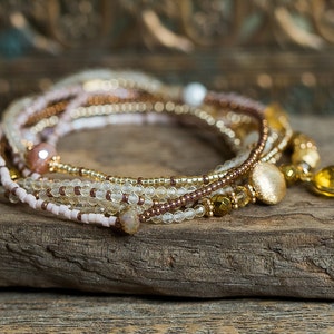 May include: A multi-strand beaded bracelet with a gold charm and a light brown, pink, and clear color scheme.