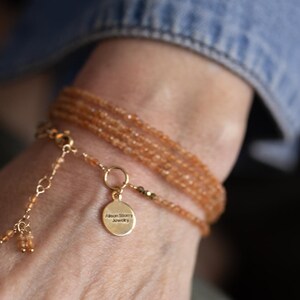 Op de afbeelding: Een meerstrengs armband met kleine, oranje-bruine kralen. De armband heeft een goudkleurige sluiting en een kleine, ronde bedel met de tekst "Alison Story Jewelry". De armband wordt om een pols gedragen, met een spijkerjas mouw op de achtergrond.
