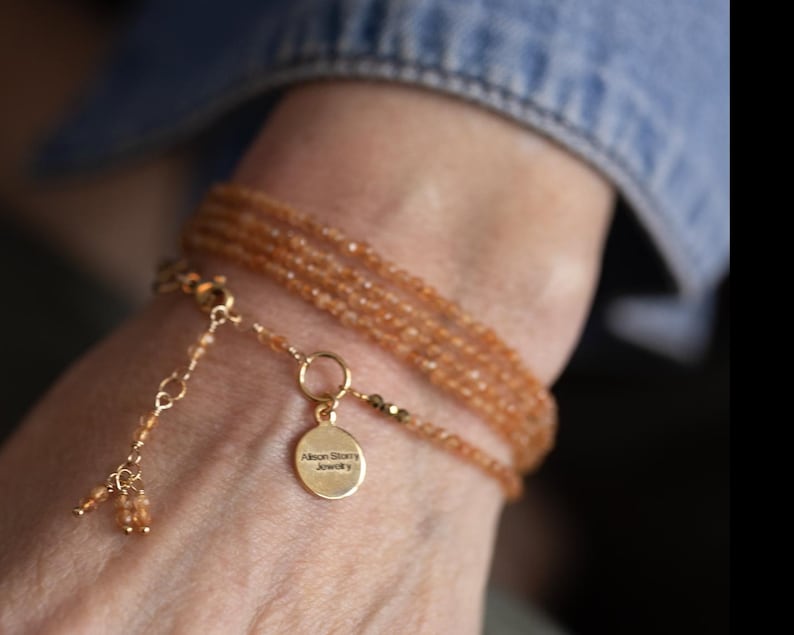 Op de afbeelding: Een meerstrengs armband met kleine, oranje-bruine kralen. De armband heeft een goudkleurige sluiting en een kleine, ronde bedel met de tekst "Alison Story Jewelry". De armband wordt om een pols gedragen, met een spijkerjas mouw op de achtergrond.