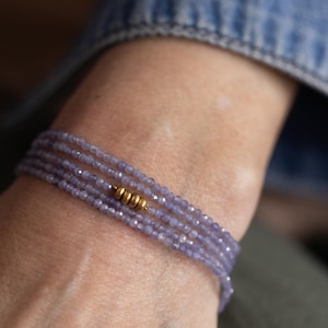 May include: A delicate bracelet featuring multiple strands of small, light purple beads. The bracelet is accented with gold-colored cylindrical beads. The jewelry is worn on a wrist, with a denim jacket sleeve visible in the background.