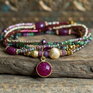 Puede incluir: Pulsera de m&uacute;ltiples hilos con un colgante de oro con una gran piedra de rub&iacute;. La pulsera est&aacute; hecha con peque&ntilde;as cuentas en tonos rosa, verde y dorado.