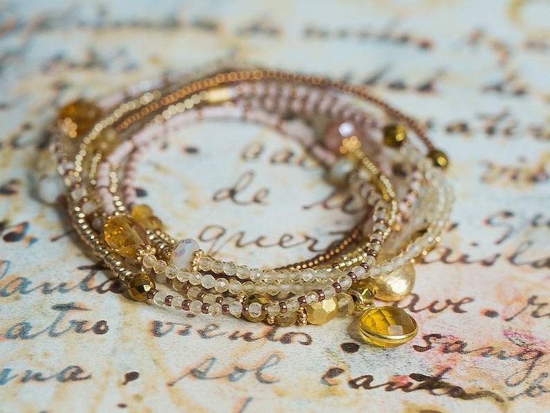 May include: A multi-strand bracelet with gold, brown, and white beads. The bracelet has a gold charm with a clear stone.