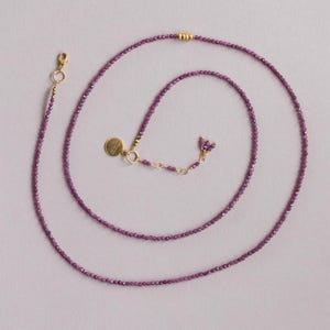 May include: A fine necklace composed of small, faceted, purple beads. It has a gold-coloured clasp and a small, round charm. The beads are a deep purple hue, and the necklace is shown against a pale background. The necklace is approximately 40 cm long.