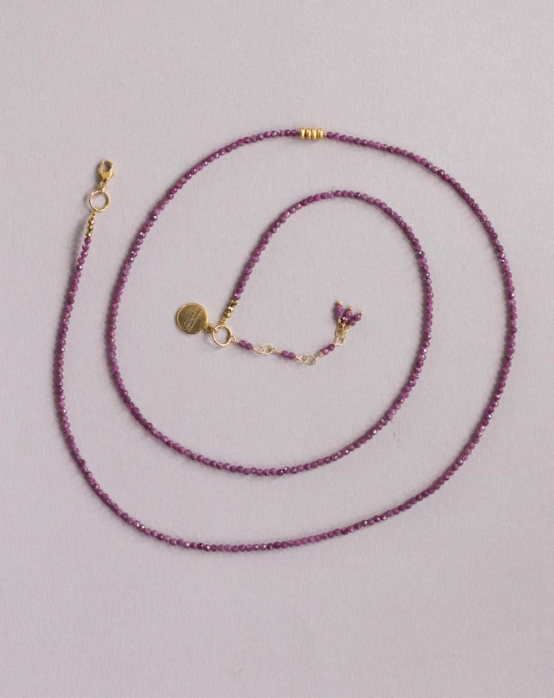 May include: A fine necklace composed of small, faceted, purple beads. It has a gold-coloured clasp and a small, round charm. The beads are a deep purple hue, and the necklace is shown against a pale background. The necklace is approximately 40 cm long.
