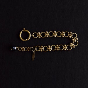 Puede incluir: Una pulsera dorada con un cierre circular y una serie de eslabones interconectados. La pulsera presenta una peque&ntilde;a perla de color oscuro en un extremo y una peque&ntilde;a etiqueta. La joya se exhibe sobre un fondo negro.
