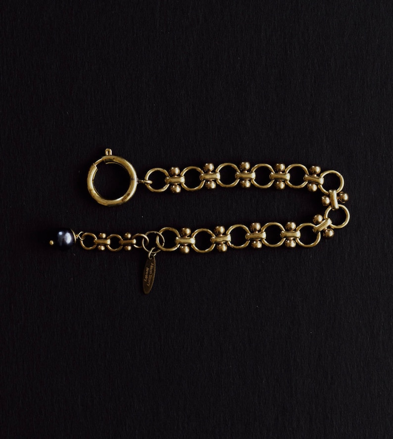 Puede incluir: Una pulsera dorada con un cierre circular y una serie de eslabones interconectados. La pulsera presenta una peque&ntilde;a perla de color oscuro en un extremo y una peque&ntilde;a etiqueta. La joya se exhibe sobre un fondo negro.