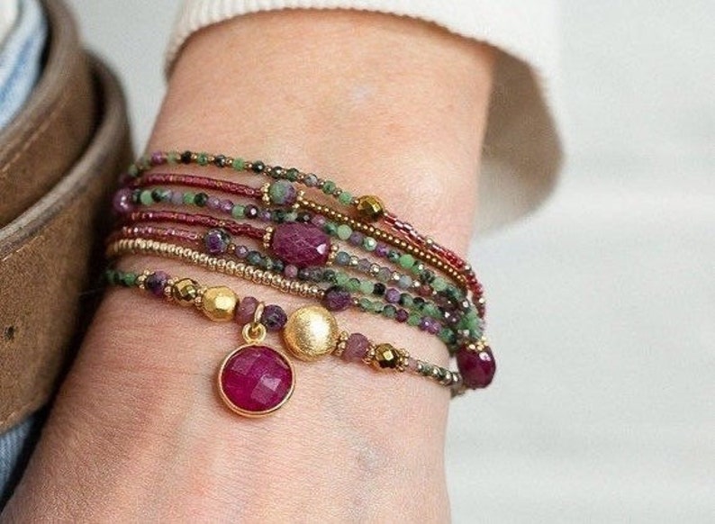 Puede incluir: Un conjunto de pulseras con cuentas rojas, verdes y doradas. Las pulseras tienen un dije con una piedra roja grande.