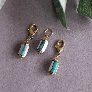 May include: Three decorative charms with gold-colored clasps and accents. Each charm features a cylindrical design with white, turquoise, and pink segments. The charms are arranged on a gray surface, with a green leaf in the background.