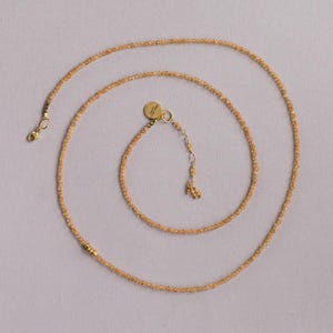 Op de afbeelding: Een delicate ketting met kleine, ronde, amberkleurige kralen. De ketting heeft een goudkleurige sluiting en een kleine, ronde bedel. De kralen zijn geregen om een lang, elegant sieraad te cre&euml;ren.