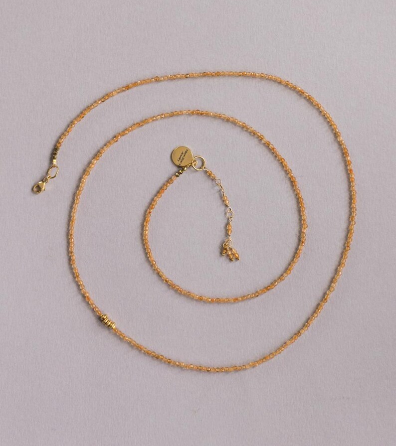 Op de afbeelding: Een delicate ketting met kleine, ronde, amberkleurige kralen. De ketting heeft een goudkleurige sluiting en een kleine, ronde bedel. De kralen zijn geregen om een lang, elegant sieraad te cre&euml;ren.
