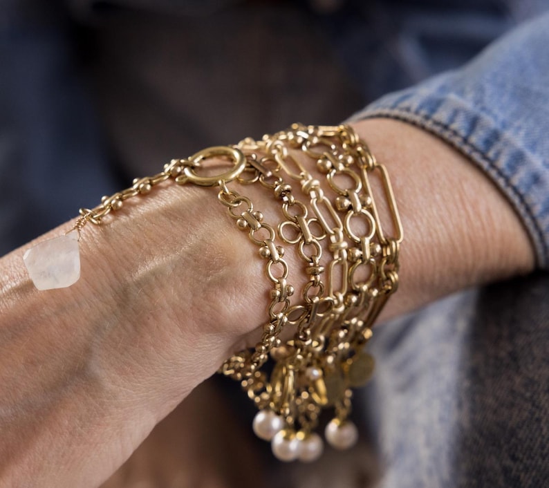 May include: A stack of gold chain bracelets with a white stone charm and pearl accents.