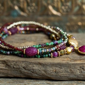 Puede incluir: Un brazalete de m&uacute;ltiples hilos con cuentas rosas, verdes y doradas. El brazalete tiene un gran colgante de piedra preciosa rosa.