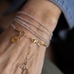K&ouml;nnte beinhalten: Ein zartes Wickelarmband mit mehreren Str&auml;ngen kleiner, hellfarbiger Perlen. Das Armband enth&auml;lt goldfarbene Akzente, einen Verschluss und einen kleinen, goldfarbenen Anh&auml;nger mit Text. Das Armband ist um ein Handgelenk gewickelt.