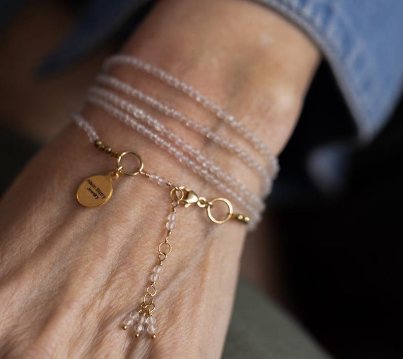 K&ouml;nnte beinhalten: Ein zartes Wickelarmband mit mehreren Str&auml;ngen kleiner, hellfarbiger Perlen. Das Armband enth&auml;lt goldfarbene Akzente, einen Verschluss und einen kleinen, goldfarbenen Anh&auml;nger mit Text. Das Armband ist um ein Handgelenk gewickelt.