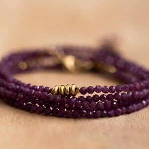 May include: A multi-strand bracelet with small, faceted, deep purple beads. The bracelet has gold-coloured metal beads and clasps. The jewellery is displayed on a light brown wooden surface.