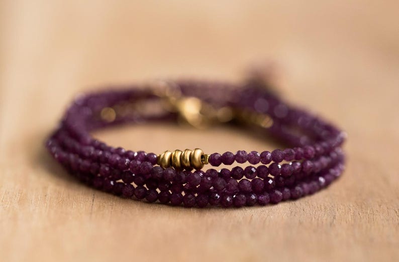 May include: A multi-strand bracelet with small, faceted, deep purple beads. The bracelet has gold-coloured metal beads and clasps. The jewellery is displayed on a light brown wooden surface.