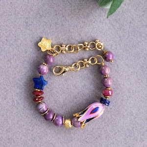 May include: A purple and gold beaded bracelet with a pink and blue flower charm, two blue star charms, and a gold star charm. The bracelet has a gold chain clasp.