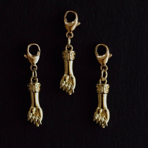 May include: Three gold-colored hand charms with lobster claw clasps. Each charm features a detailed hand design with an ornate cuff. The charms are suspended from the clasps, creating a dangling effect.