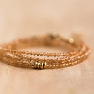 Op de afbeelding: Een delicate, meerstrengs armband met kleine, ronde, amberkleurige kralen. De armband is geaccentueerd met goudkleurige metalen kralen en sluitingen. De sieraden worden gepresenteerd op een lichtgekleurd houten oppervlak.