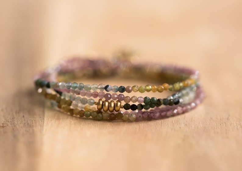 Pu&ograve; includere: Un insieme di braccialetti di perline nei toni del rosa, verde, giallo e nero. I braccialetti sono fatti di piccole perline rotonde e presentano dettagli metallici color oro. Sono esposti su una superficie di legno.