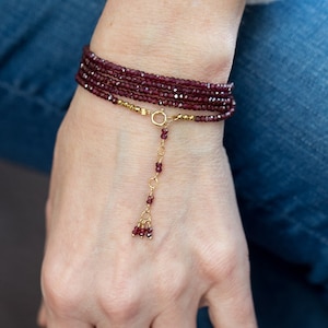 Op de afbeelding: Een meerstrengs armband met een hangende bedel. De armband is gemaakt van kleine, donkerrode kralen en een gouden ketting. De bedel is een cluster van kleine, donkerrode kralen.