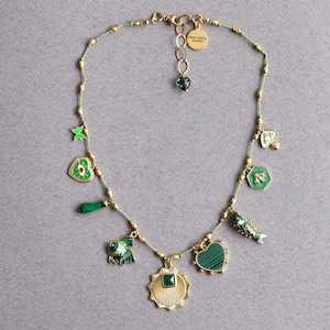 May include: A green and gold necklace with a variety of charms, including a heart, a fish, a bee, a mushroom, and a butterfly. The necklace is made of a thin, green cord and has a gold clasp. The charms are all made of metal and have a green enamel finish. The necklace is labeled "Alice Story Jewelry".