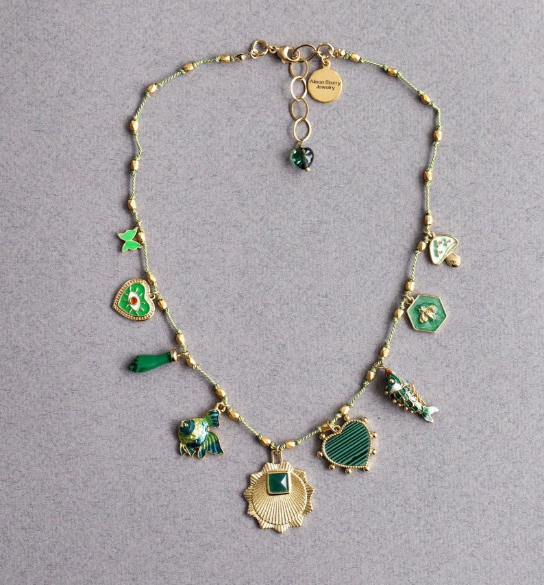 May include: A green and gold necklace with a variety of charms, including a heart, a fish, a bee, a mushroom, and a butterfly. The necklace is made of a thin, green cord and has a gold clasp. The charms are all made of metal and have a green enamel finish. The necklace is labeled "Alice Story Jewelry".