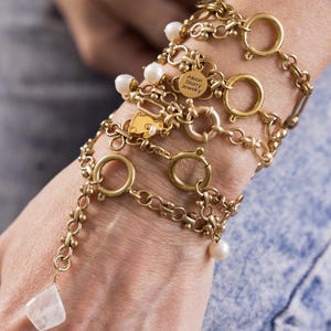 May include: A gold chain bracelet with multiple charms, including a round charm with the text "Alison Story Jewelry". The bracelet also features small pearl accents.