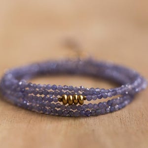 May include: A delicate wrap bracelet featuring multiple strands of small, faceted, lavender-colored beads. The bracelet is accented with gold-colored metal beads. The jewelry is displayed on a light brown wooden surface.
