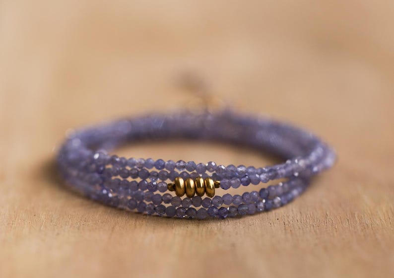May include: A delicate wrap bracelet featuring multiple strands of small, faceted, lavender-colored beads. The bracelet is accented with gold-colored metal beads. The jewelry is displayed on a light brown wooden surface.