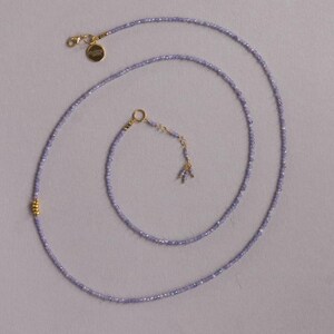 May include: Delicate lavender beaded necklace with gold-colored accents. The necklace features a small gold clasp and a gold charm. The beads are small and evenly spaced, creating a subtle, elegant look. The necklace is designed to be worn around the neck.