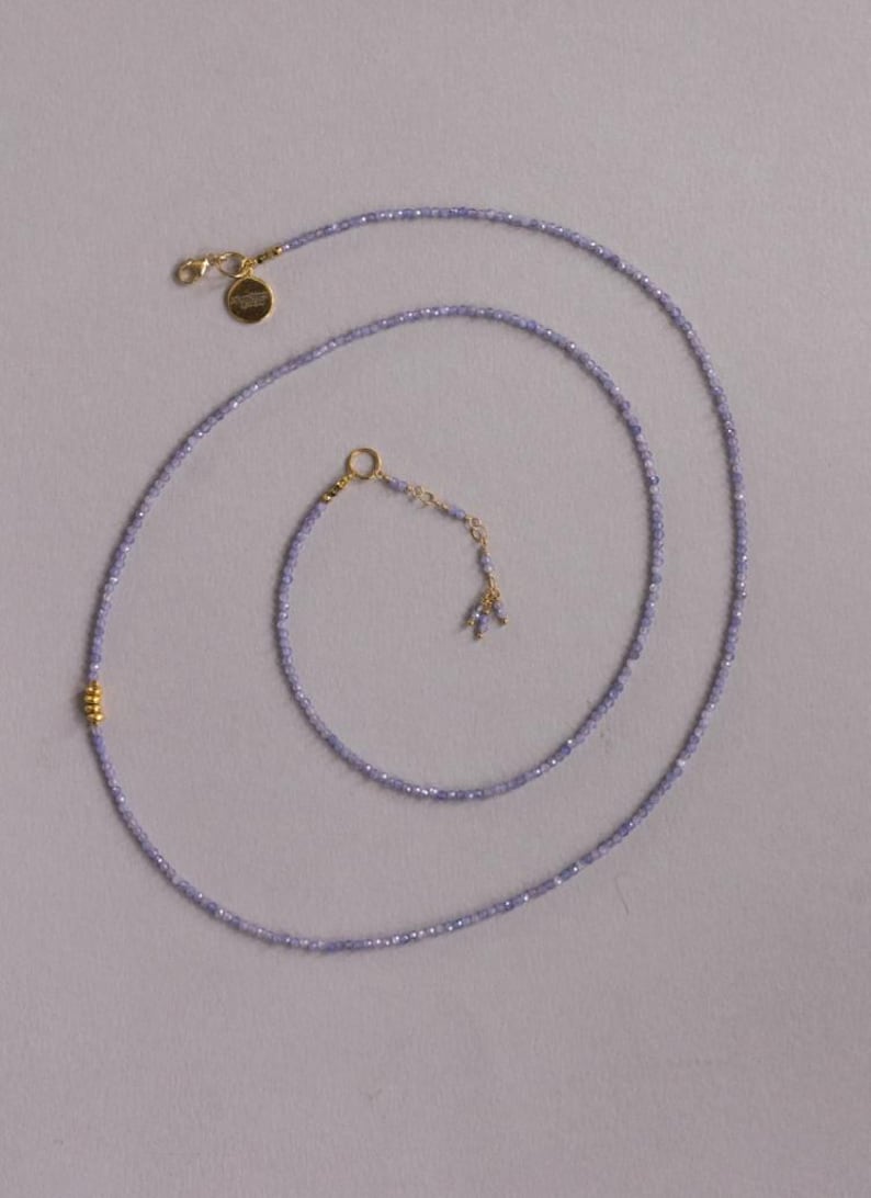 May include: Delicate lavender beaded necklace with gold-colored accents. The necklace features a small gold clasp and a gold charm. The beads are small and evenly spaced, creating a subtle, elegant look. The necklace is designed to be worn around the neck.