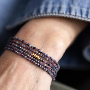 May include: A multi-strand bracelet featuring small, round, dark blue and purple beads. The bracelet has a gold-colored cylindrical accent. The bracelet is worn on a wrist, with a denim jacket in the background.