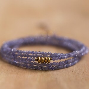 May include: A delicate wrap bracelet featuring multiple strands of small, faceted, lavender-colored beads. The bracelet is accented with gold-colored metal beads. The jewelry is displayed on a light brown wooden surface.