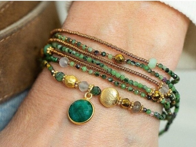K&ouml;nnte beinhalten: Ein mehrreihiges Armband mit gr&uuml;nen und braunen Perlen, goldenen Akzenten und einem gro&szlig;en gr&uuml;nen Edelstein-Anh&auml;nger.
