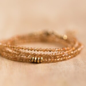 Op de afbeelding: Een delicate, meerstrengs armband met kleine, ronde, amberkleurige kralen. De armband is geaccentueerd met drie goudkleurige cilindrische kralen en wordt gepresenteerd op een lichtgekleurd houten oppervlak.