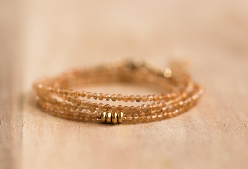 Op de afbeelding: Een delicate, meerstrengs armband met kleine, ronde, amberkleurige kralen. De armband is geaccentueerd met drie goudkleurige cilindrische kralen en wordt gepresenteerd op een lichtgekleurd houten oppervlak.